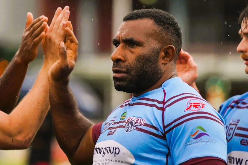 A rugby league player from Papua New Guinea wearing a light blue jersey with maroon horizontal stripes high fiving teammates.