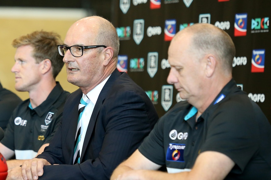 Four men sitting for a press conference, a bald man in the centre wearing a suit