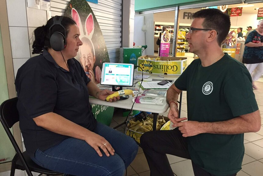 Naomi, wearing headphones, and Neil sitting down at a shopping centre with boxes of bananas in the background