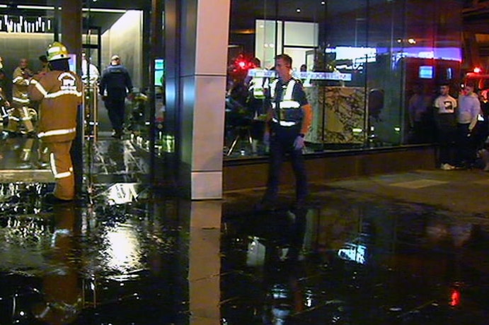 Water floods out the front doors of the Experience Bella Hotel Apartments with a fireman and police officer on the scene.