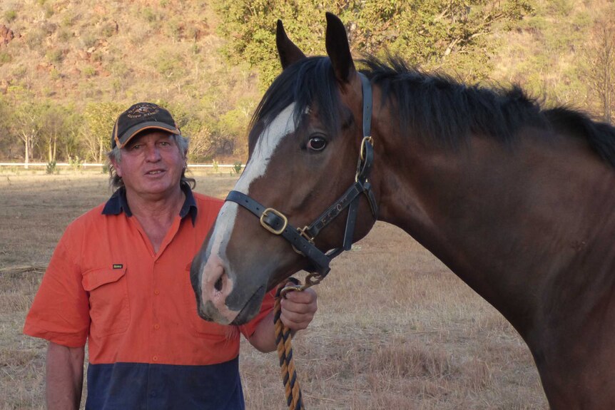 A man in an orange working shirt standing next to a racehorse with hills in the background.