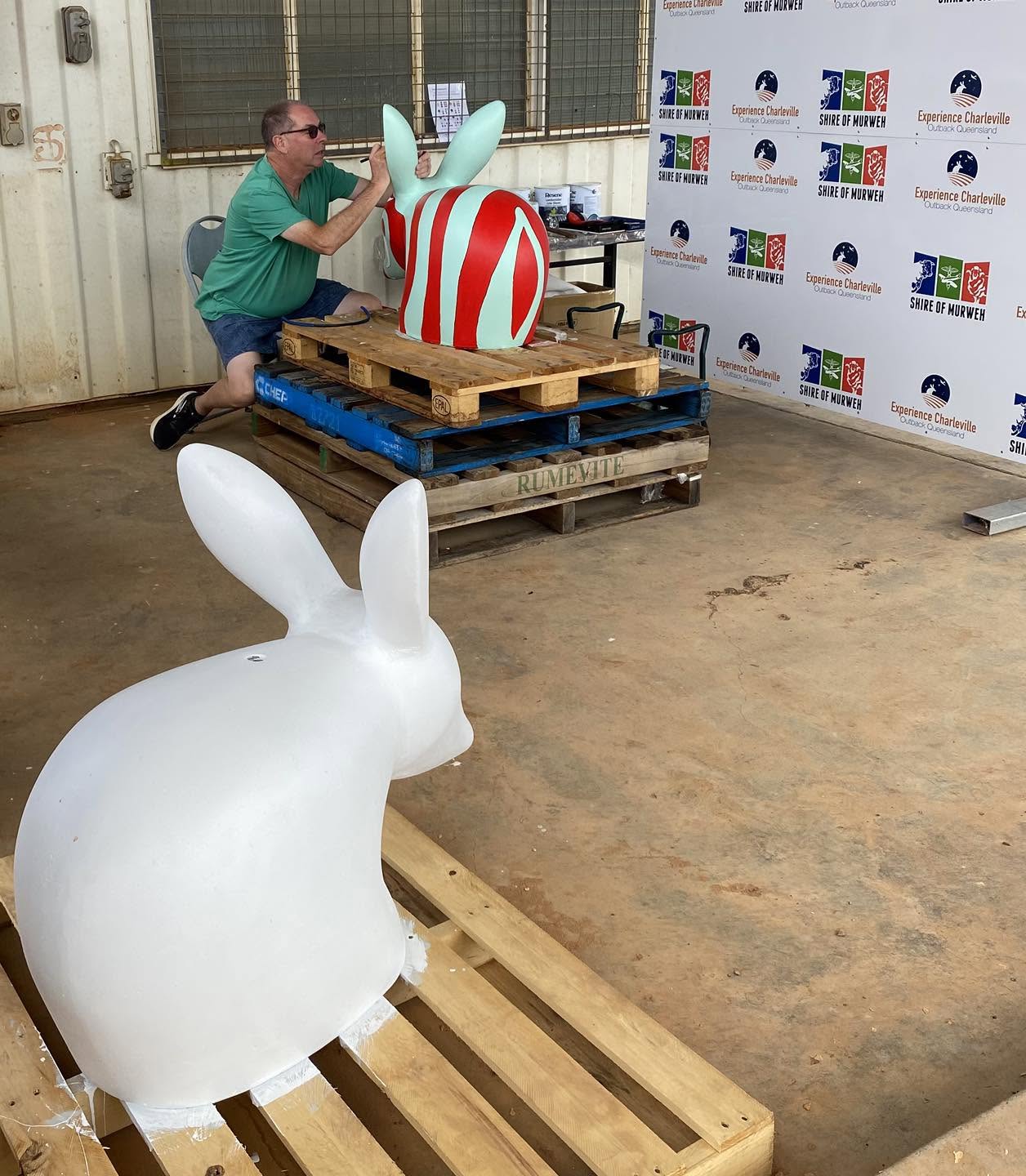 a man paints a cement bilby on a wooden pallet, with an unpainted bilby in the foreground