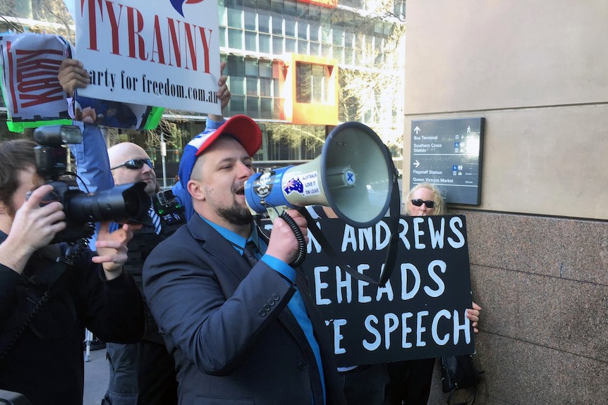 Neil Erikson yells into a megaphone while holding a "Dan Andrews beheads free speech" sign outside court.
