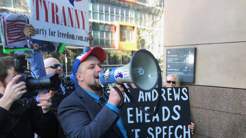 The UPF's Neil Erikson yells into a megaphone while holding a "Dan Andrews beheads free speech" sign outside court.