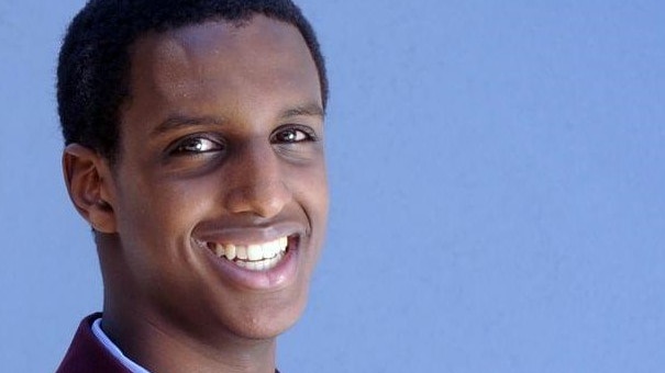 Raghe Mohammed Abdi smiles a the camera. He wears a school uniform consisting of a maroon jacket and tie, and a blue shirt.