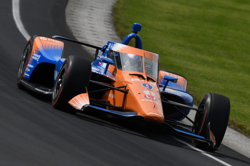 An orange and blue IndyCar drives around a banked corner
