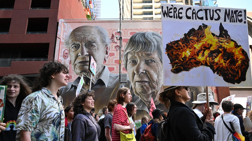 Protesters march along a street with one carrying a banner stating 'We're cactus mate' against an image of Australia burning