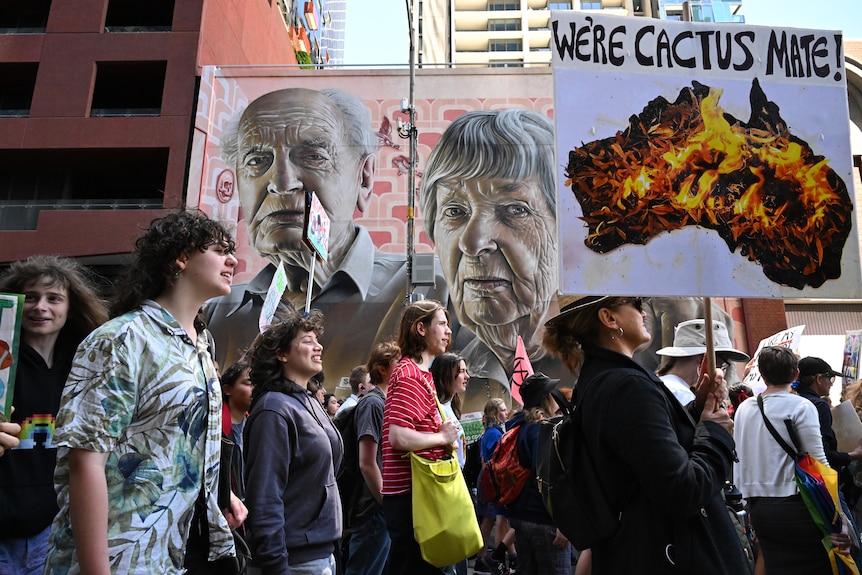 Protesters march along a street with one carrying a banner stating 'We're cactus mate' against an image of Australia burning