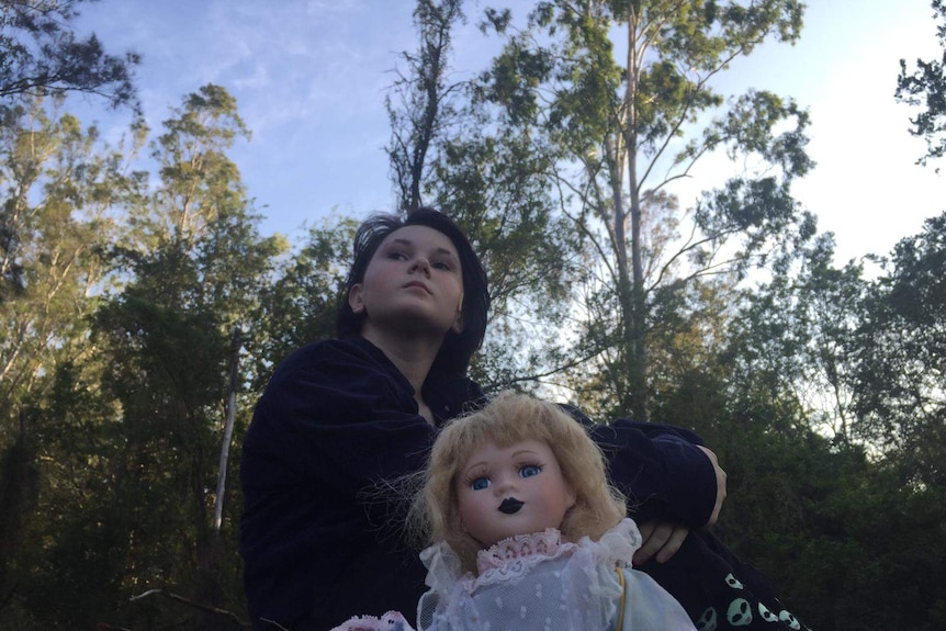 A teenage girl in dark clothes sits on grass with a doll, looking into the distance.