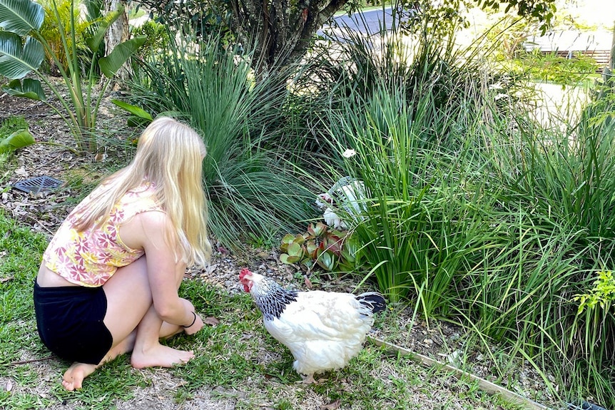 A girl with long blonde hair crouches near some chickens in a garden