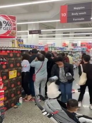 A crowd of people grouped at a small portion of a shopping centre aisle, grabbing baby formula tins.