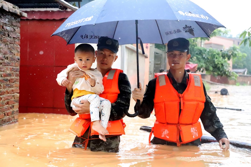 Two men in camouflage and high vis vests carry a young child through floodwater and hold an umbrella.