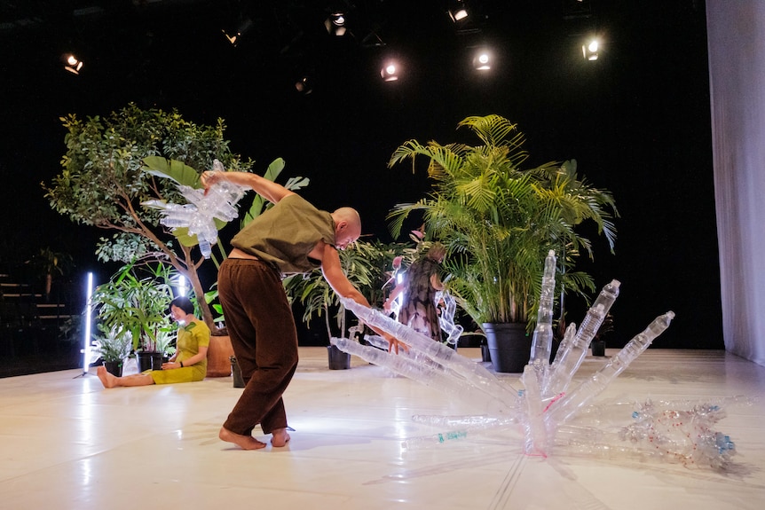 A bald man on stage holds a sculpture made of plastic bottles in one hand, reaching down to grasp a second one with the other