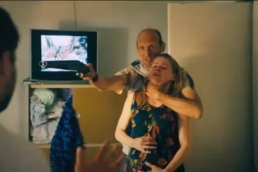 A man holds a woman around the neck and points a gun at another man