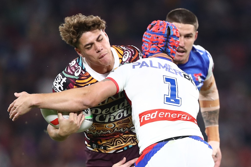 A Brisbane Broncos NRL player holds the ball as he is tackled by a Newcastle opponent.