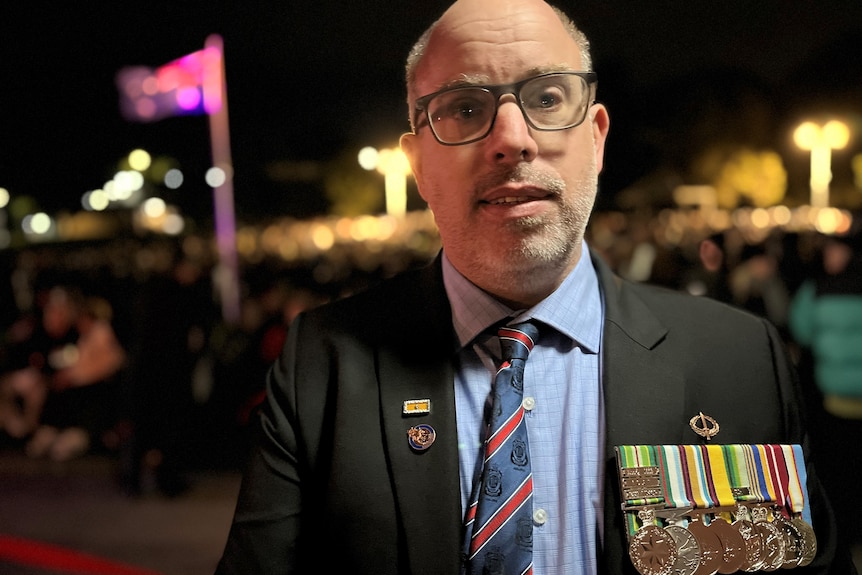 A man wearing war medals stands in front of crowd in darkness.