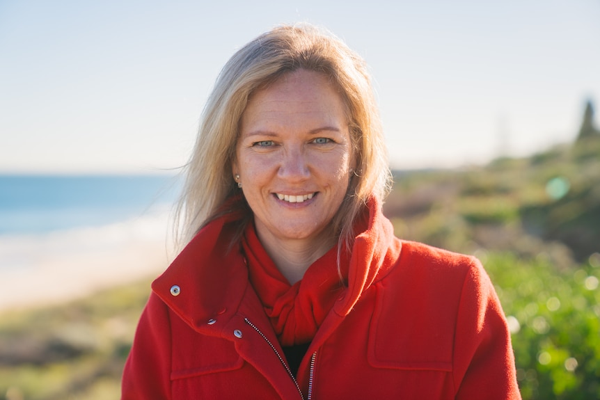 Bourby wears a bright red jacket and red neck scarfe, in a close up photo, smiling with a beach in the background.