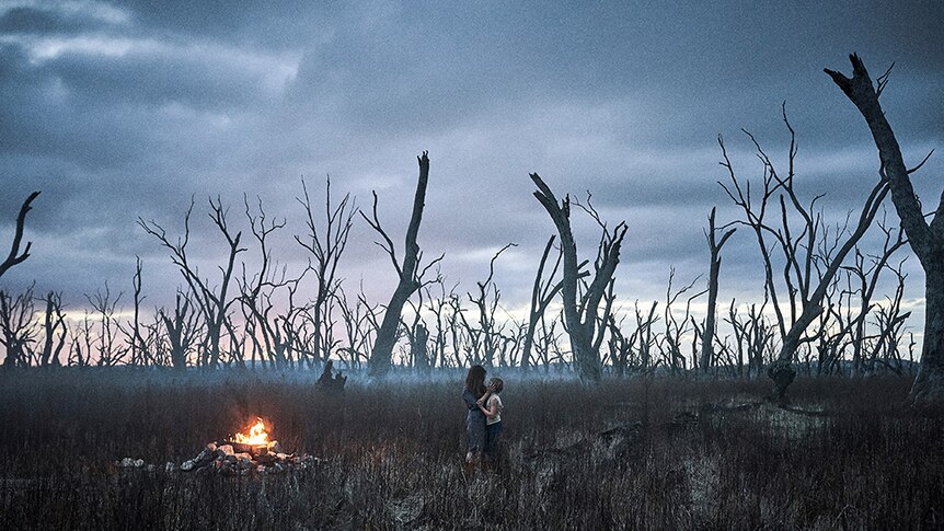A mother and son embrace near fire at sunset amongst field of dead trees on a moody and cloudy day.
