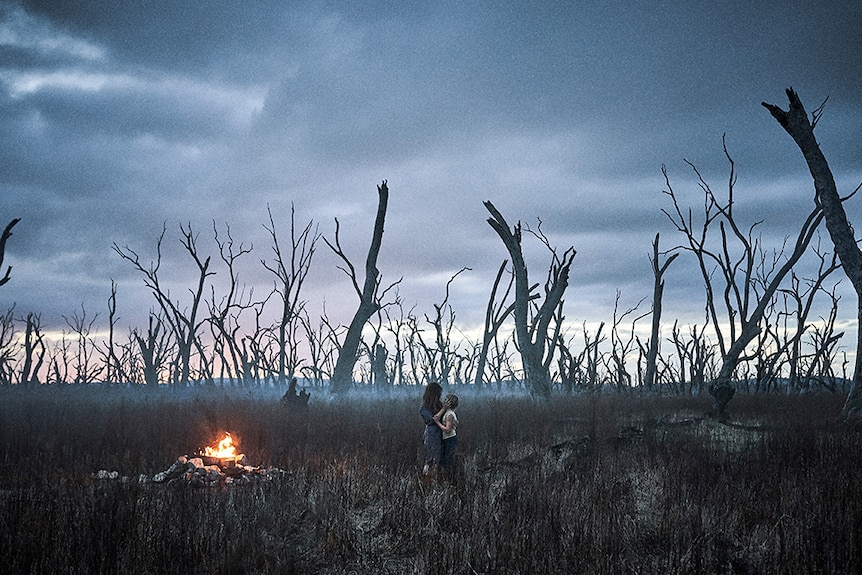 A mother and son embrace near fire at sunset amongst field of dead trees on a moody and cloudy day.