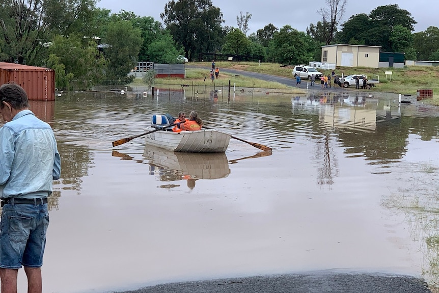 Flood water over road, two people in a small boat on the water, cars and residents in the background.