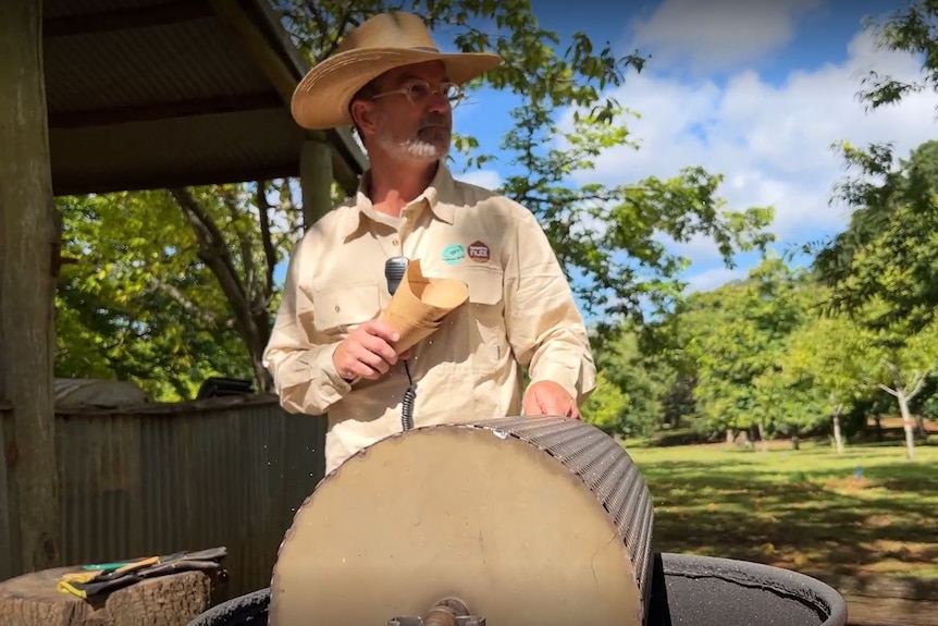 Peter Dover wears a light brown shirt and hat and turns a chestnut roaster over a fire.