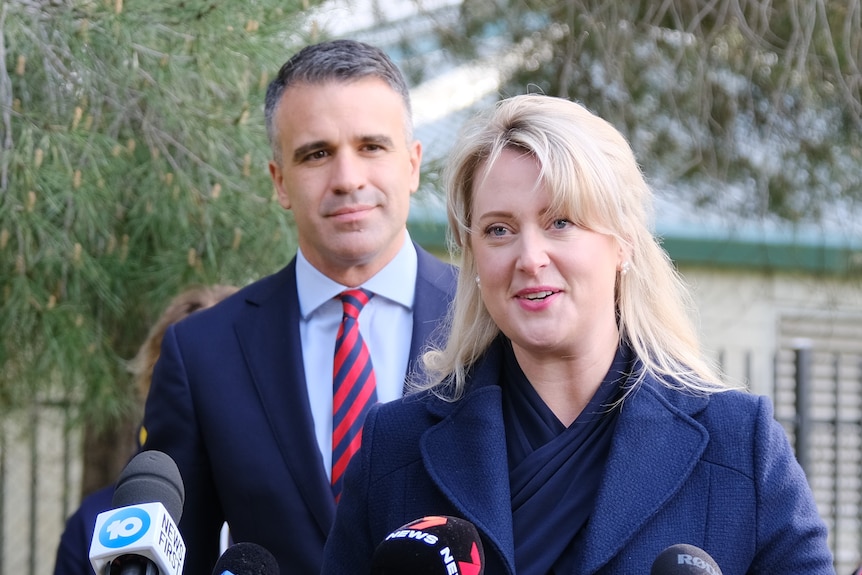 A woman wearing a dark blue coat stands in front of media microphones. A man in a blue suit stands behind her to the left