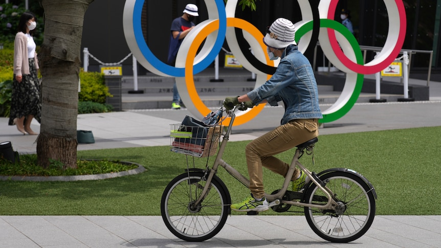 A woman wearing a hat rides in front of the Olympic rings in a park.