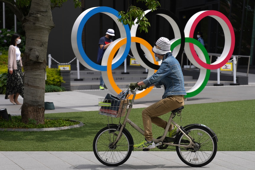 A woman wearing a hat rides in front of the Olympic rings in a park.