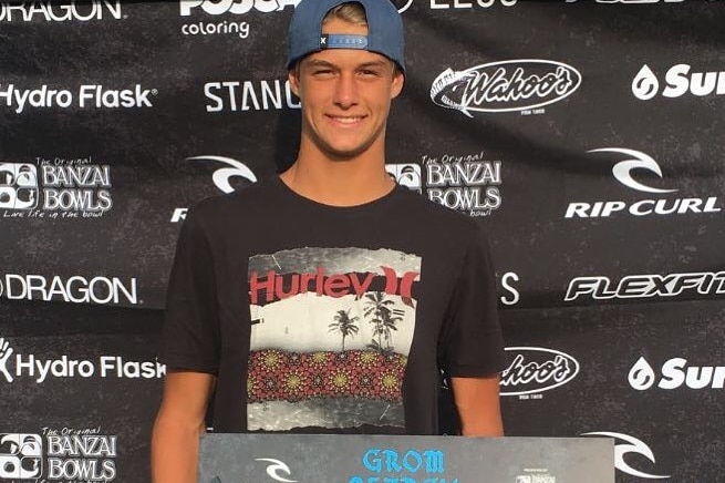 A teenager stands on a podium with a winners cheque, smiling with his hat on backwards.
