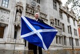 A man stands outside the supreme court holding a large Scottish flag. 