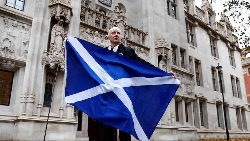 A man stands outside the supreme court holding a large Scottish flag. 