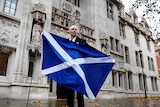 A man stands outside the supreme court holding a large Scottish flag. 