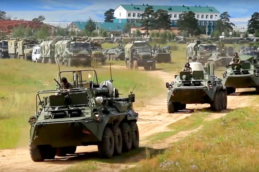 Green army tanks and troop carriers driving along a dusty round with a handful of medium sized buildings in the background