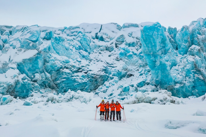 A team of young women rugged up in big red puffer jackets stand against the backdrop of a glacier