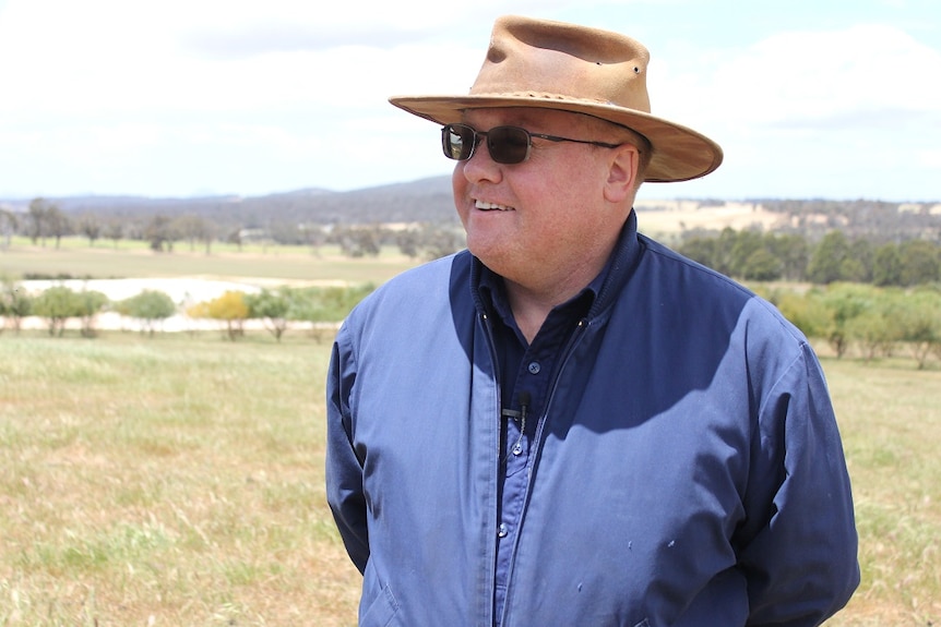 A man wearing a blue shirt, leather sunhat and sunglasses stands on farm land.
