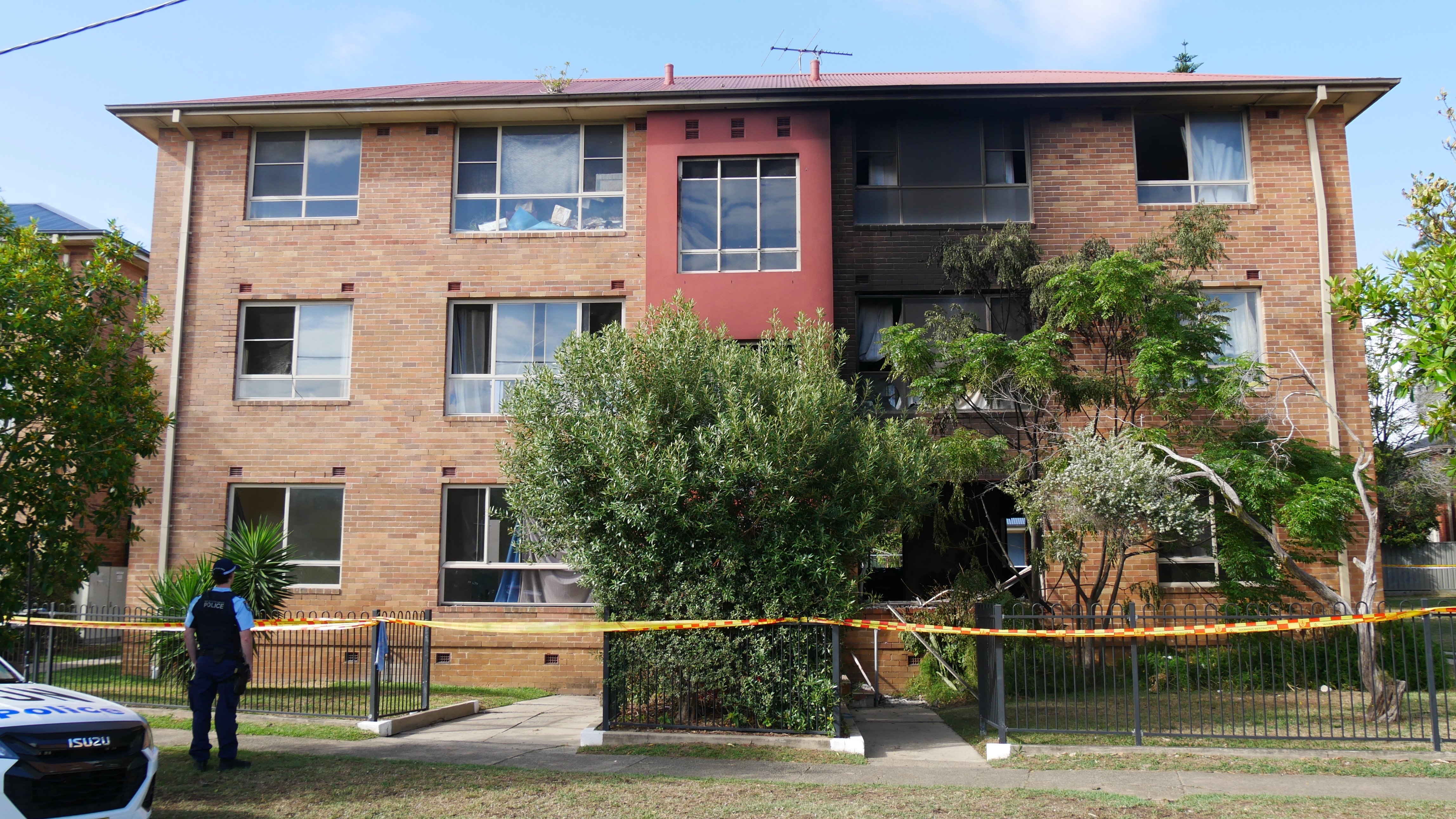Exterior no dia do bloco de unidades, queimado nos dois andares exteriores superiores, fita policial ao redor, árvores do lado de fora. 