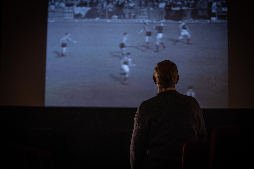 A man watches an old football game on a projector screen.