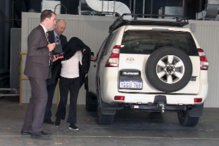 A Perth mother hides under a black jacket while being escorted by two police officers.