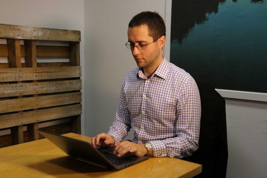 A man sits at a table typing on his laptop in front of a picture of a water landscape.