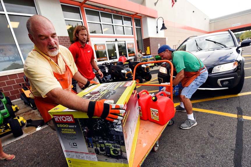 A man lifts a generator in a box on to a trolley outside a shop, while two other men load supplies behind him