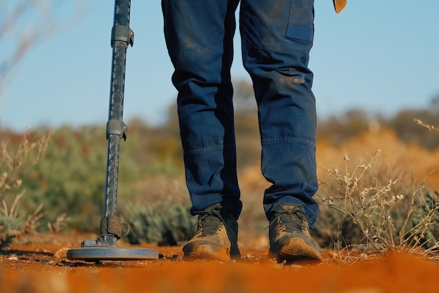 A person holds a metal detector over red dirt.