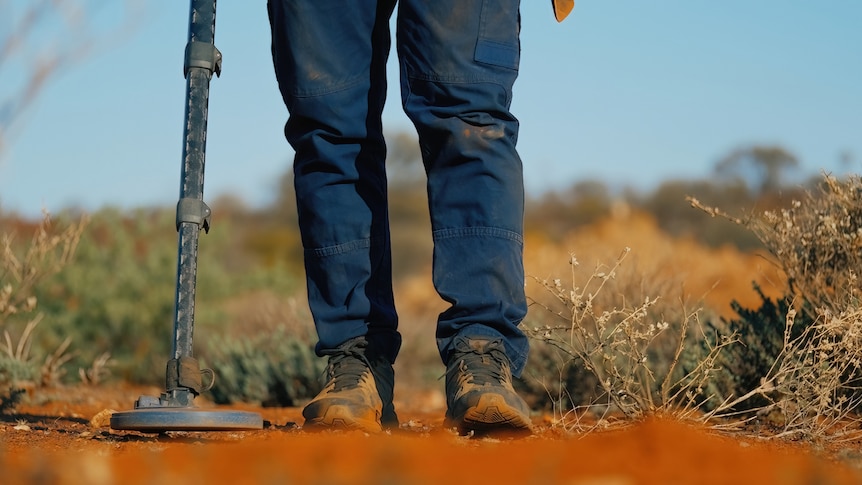 A person holds a metal detector over red dirt.