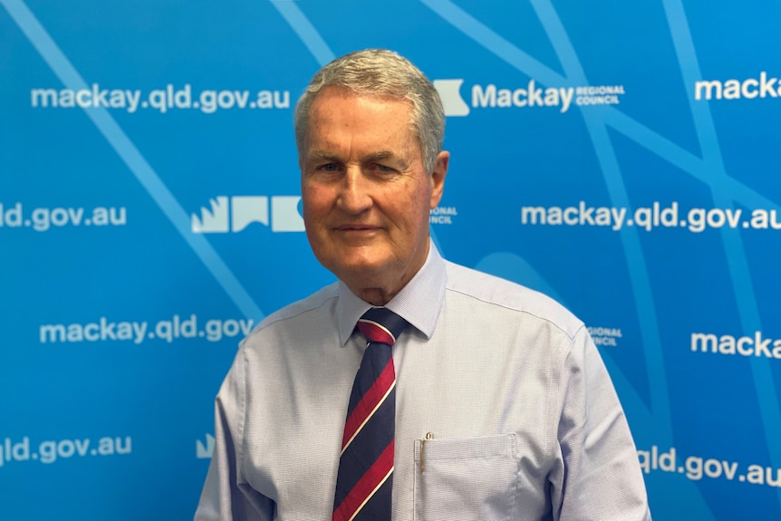 A late middle-aged man stands in front of a blue backdrop that says "Mackay Regional Council".