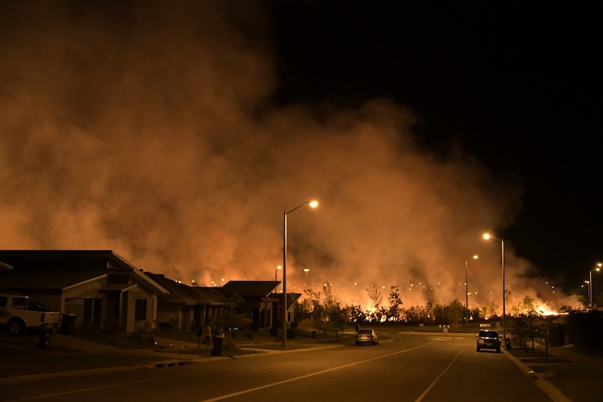 A suburban street, lit by an orange glow of scrub fires in the background