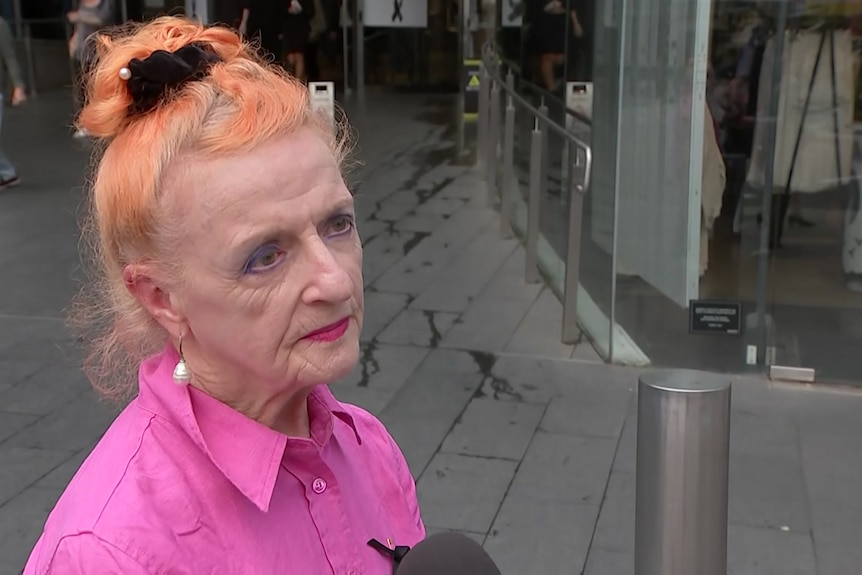 A woman in a pink shirt talks to a camera crew in front of a shopping centre entrance.
