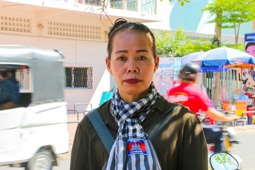A woman in a Cambodian flag scarf stands on a street corner while cars and scooters go by