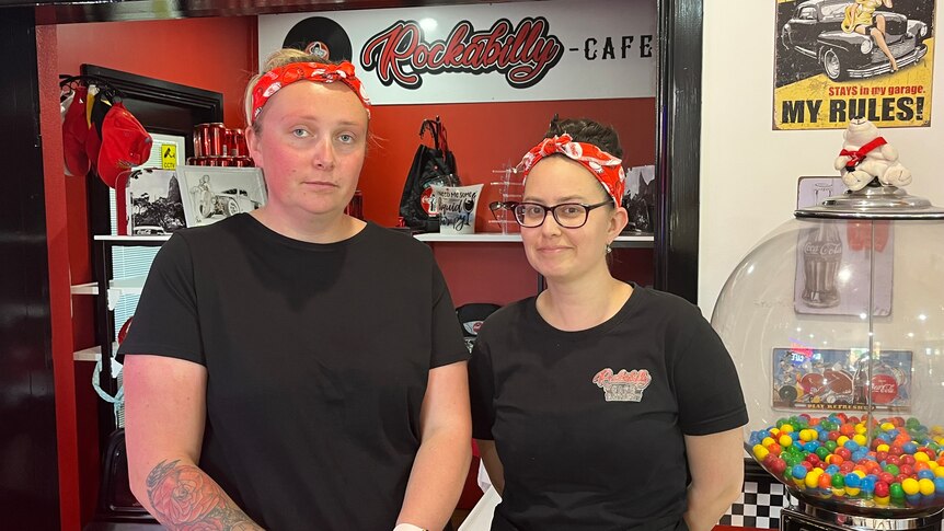 Two women standing in cafe, one smiles, both wear a red bandanna, black t-shirt one has tattoos on arm.