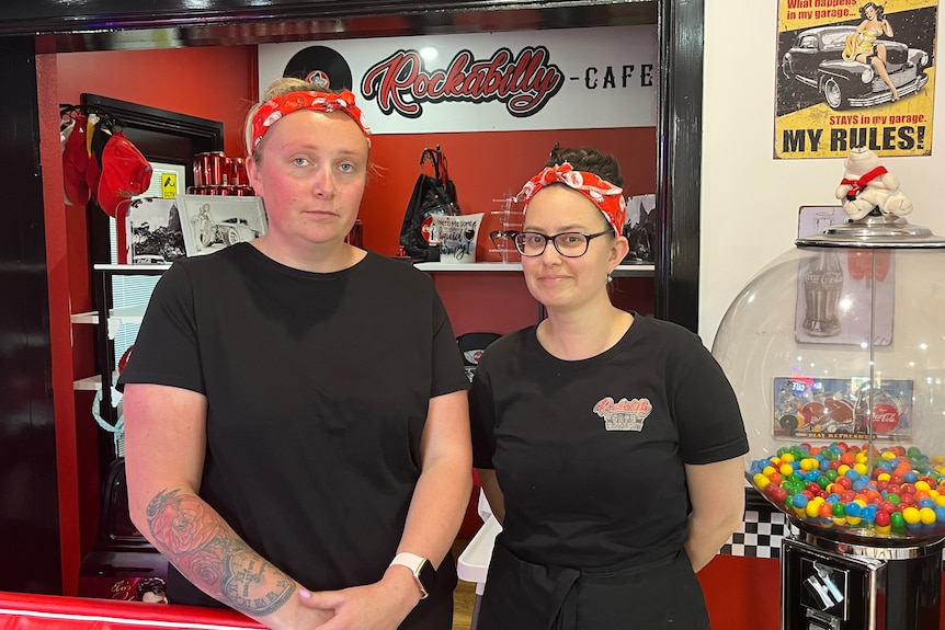 Two women standing in cafe, one smiles, both wear a red bandanna, black t-shirt one has tattoos on arm.