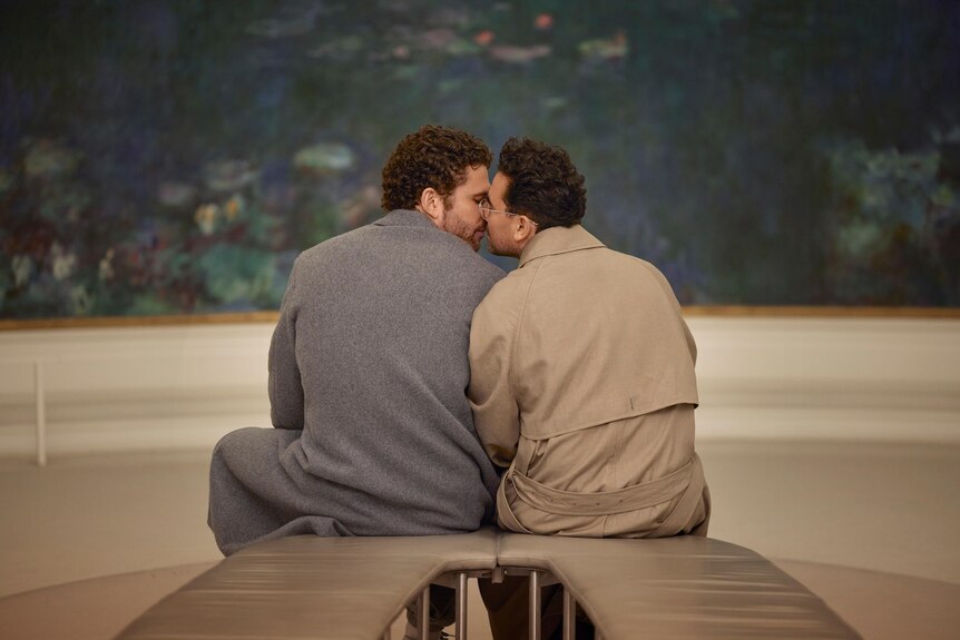 A film still of Arnaud Valois and Daniel Levy, seated kissing in an art gallery in front of an impressionist painting.
