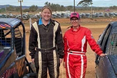 Two women in racing overals between two racecars at the dirt racing track, white bumper tyres in background